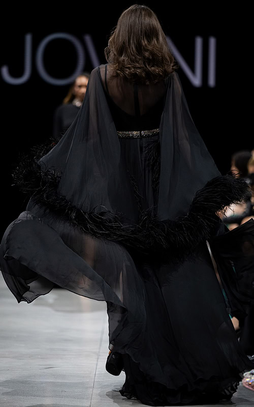 Model wearing S39276 black gown, back view with sheer feathered sleeves.